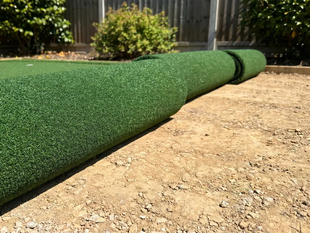 A roll of short, dense green putting-green turf partly unrolled over a compacted tan base in a backyard